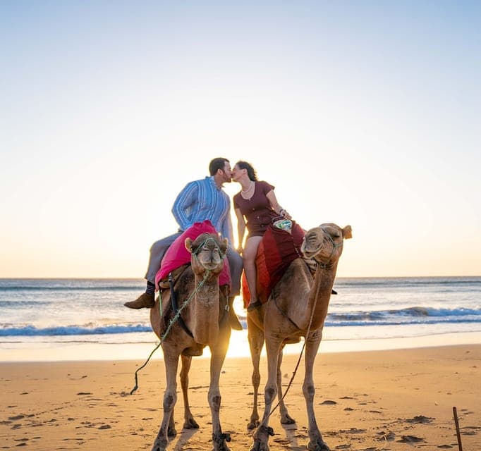Couple kissing while riding camels on a sandy beach at sunset with ocean waves.