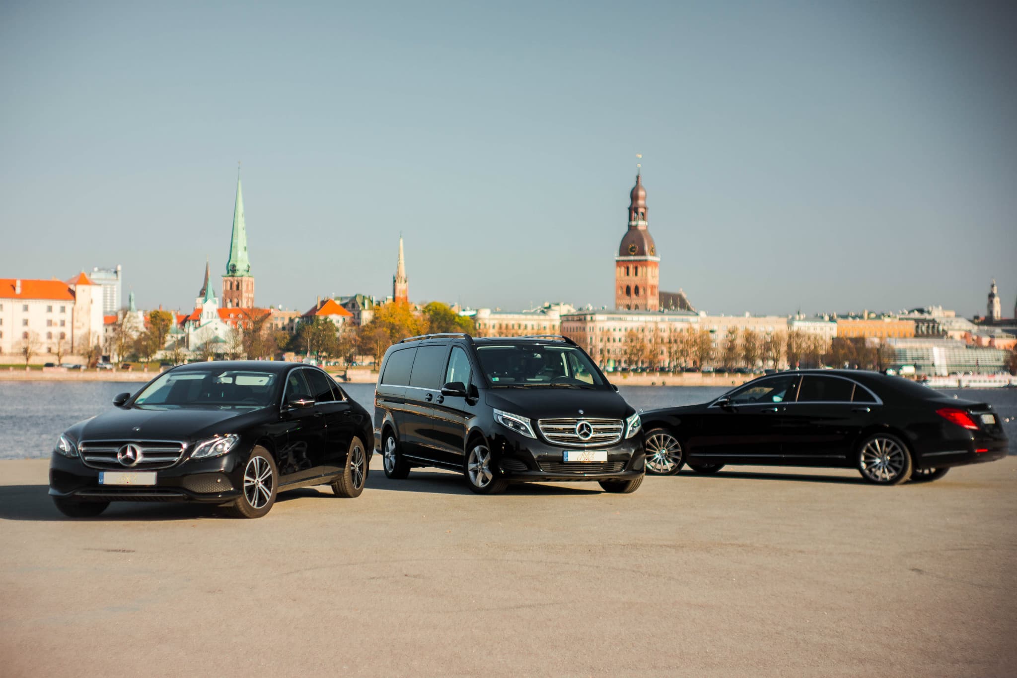 Three black Mercedes-Benz vehicles parked on a waterfront with the historic Riga city skyline.