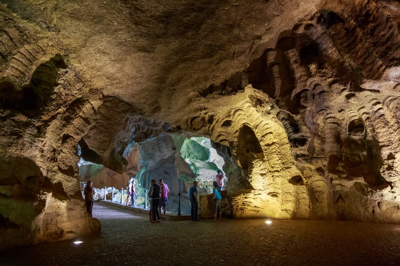 Tourists explore a large grotto with walls featuring intricate, shell-like patterns under artificial lighting.