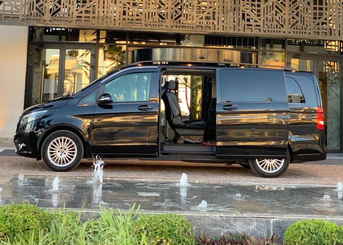 Black Mercedes-Benz van with open sliding door parked beside a fountain and modern building.