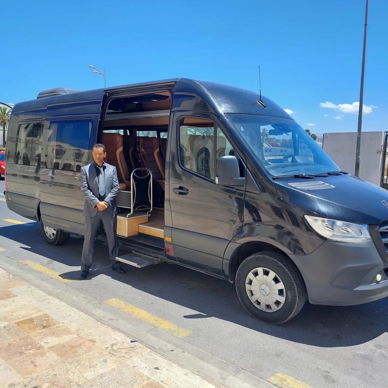 Chauffeur in a grey suit stands by a black luxury van with tan leather seats.