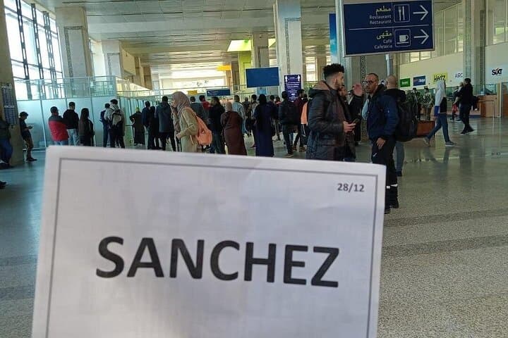 A handheld sign reading "SANCHEZ" in the foreground of a busy, sunlit airport terminal.